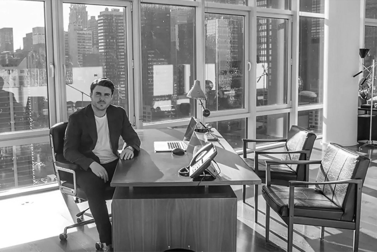 Sam Ovens sitting at his office desk in front of floor-to-ceiling windows overlooking a city skyline